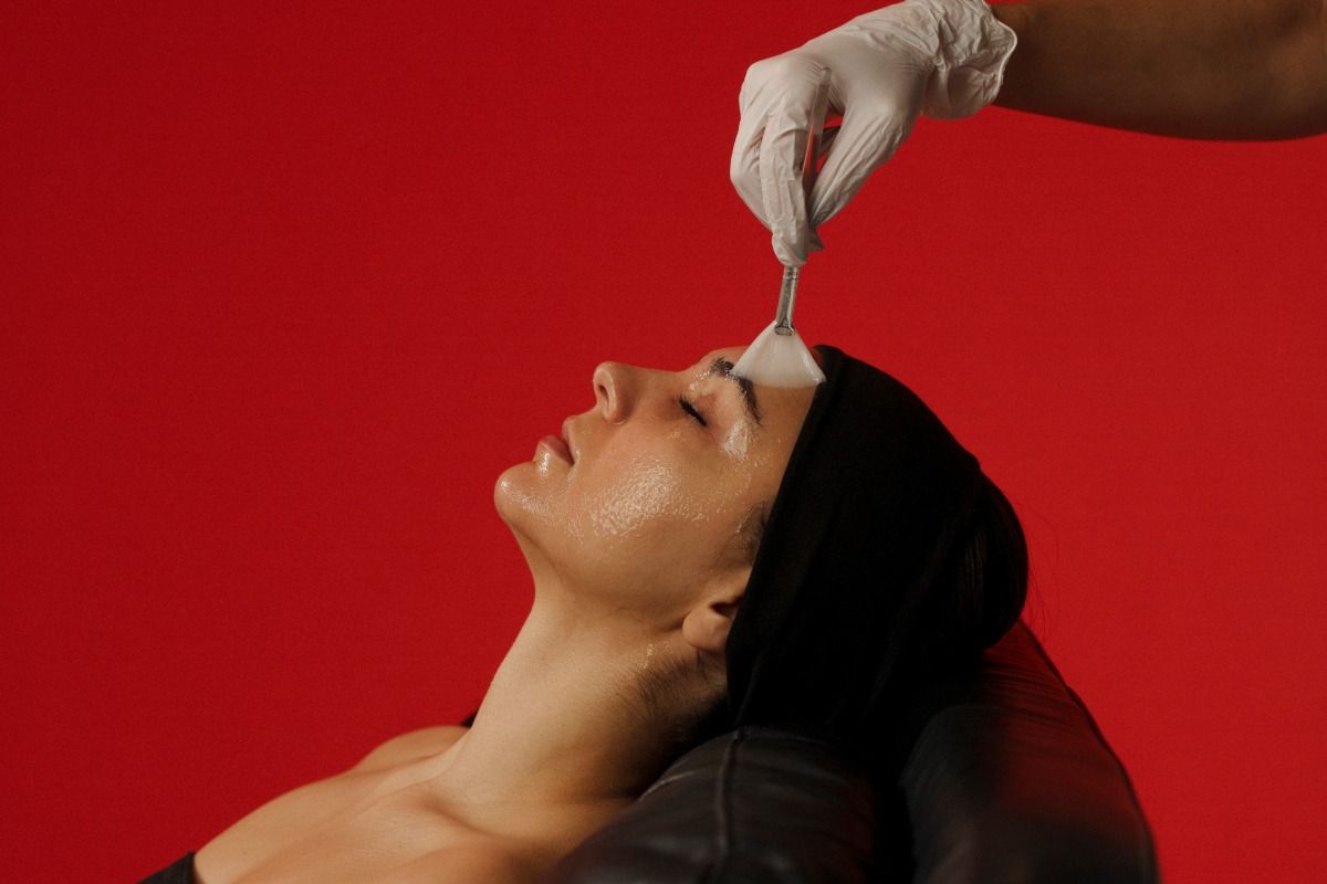 Person receiving a facial treatment with a brush on forehead, wearing a headband, against a red background.