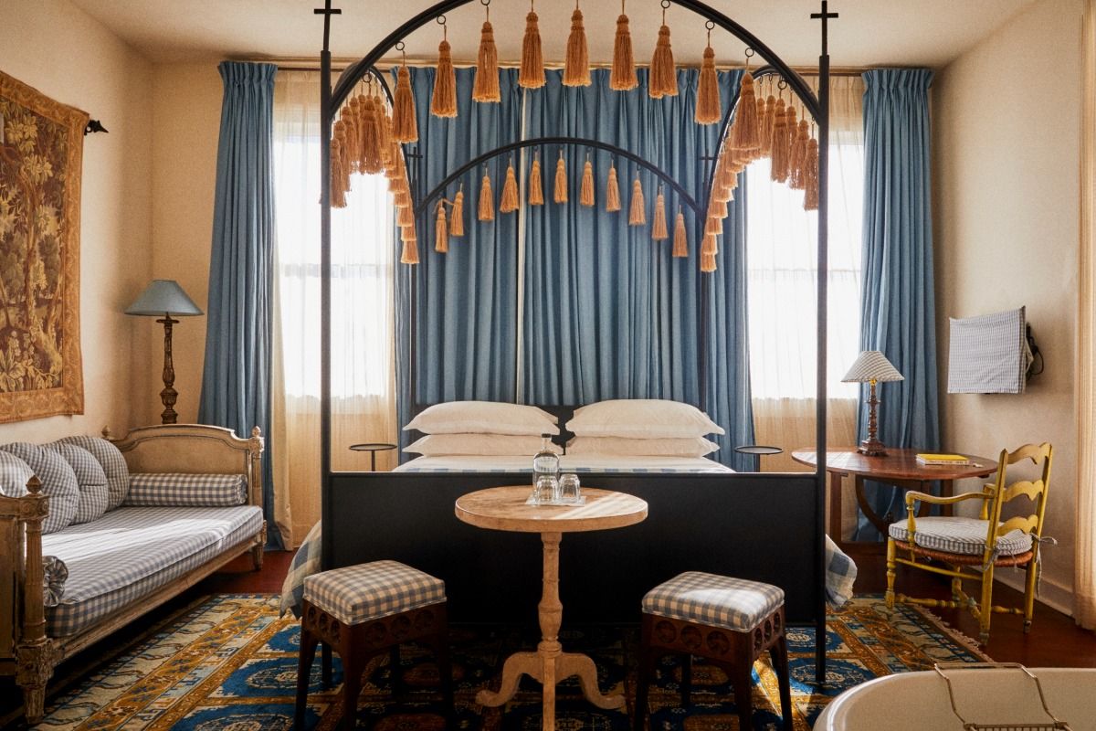 Cozy bedroom with a canopy bed, tasseled drapery, plaid furniture, wooden table, blue curtains, and a decorative rug.