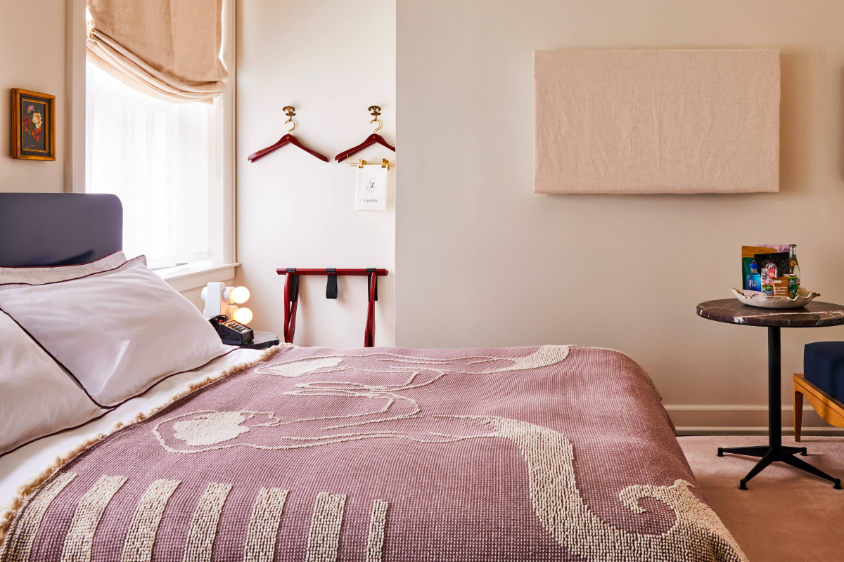 Cozy bedroom with a bed covered by a patterned blanket, wall art, and a small table with books. Natural light from a window with a blind.