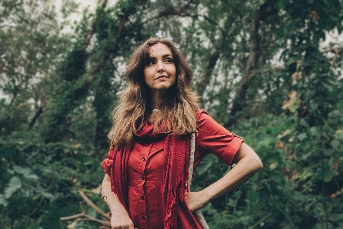 A woman in a red dress and scarf stands confidently in a lush forest, with her hands on her hips, surrounded by greenery.