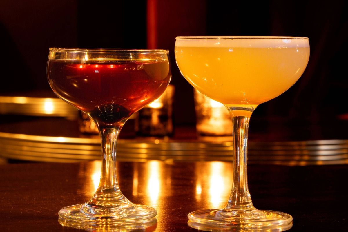 Two cocktail glasses; one with a dark red drink, the other with an amber-orange drink, set against a warm, dimly lit background.