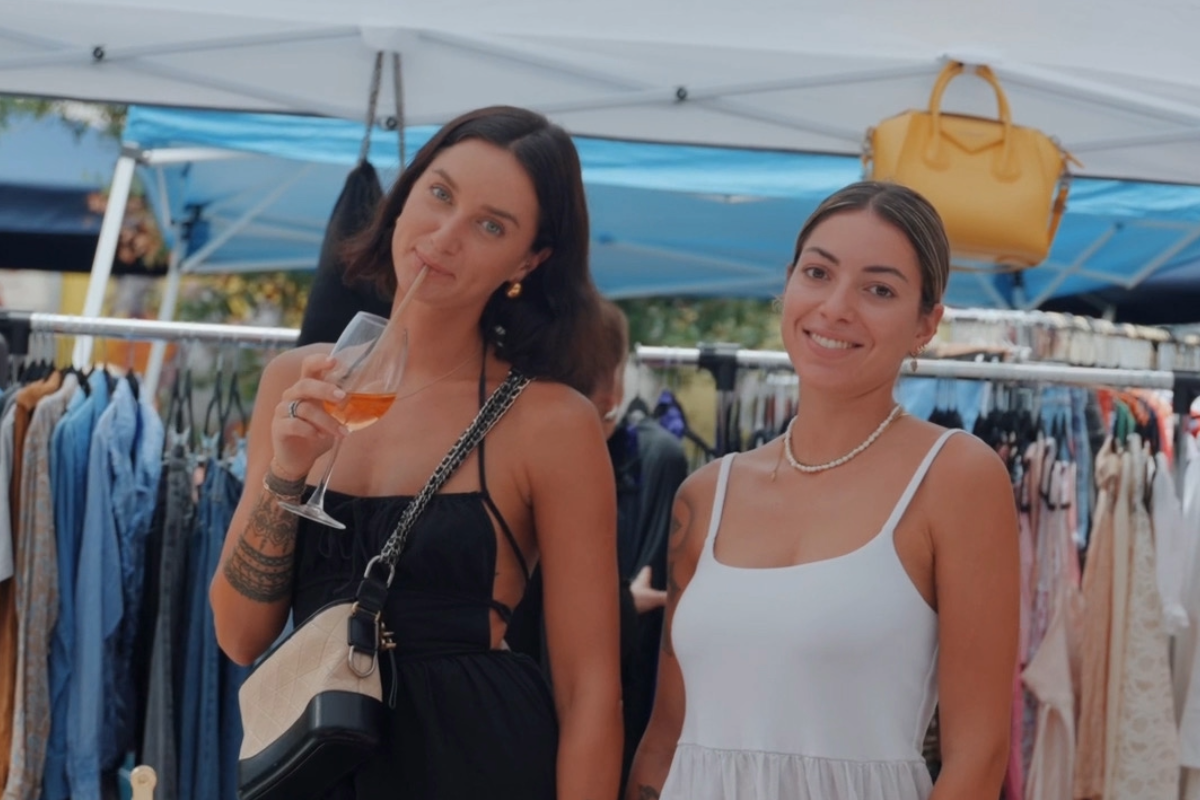 Two women at an outdoor market; one holds a drink, wearing black, the other in white smiles. Clothes and a yellow bag in the background.