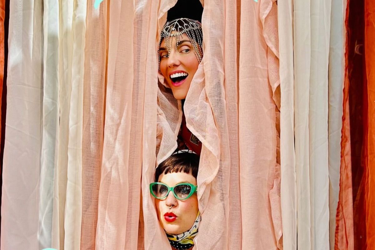 Two people peeking through pink curtains; one smiling with a jeweled headpiece, the other wearing green glasses and a headscarf, both expressing surprise.