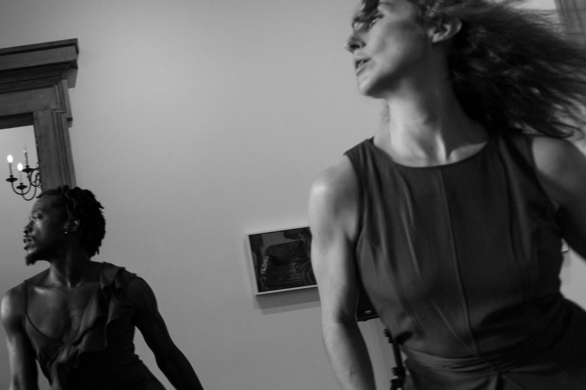Two people in motion, captured in black and white, indoors with a framed picture and a chandelier in the background.