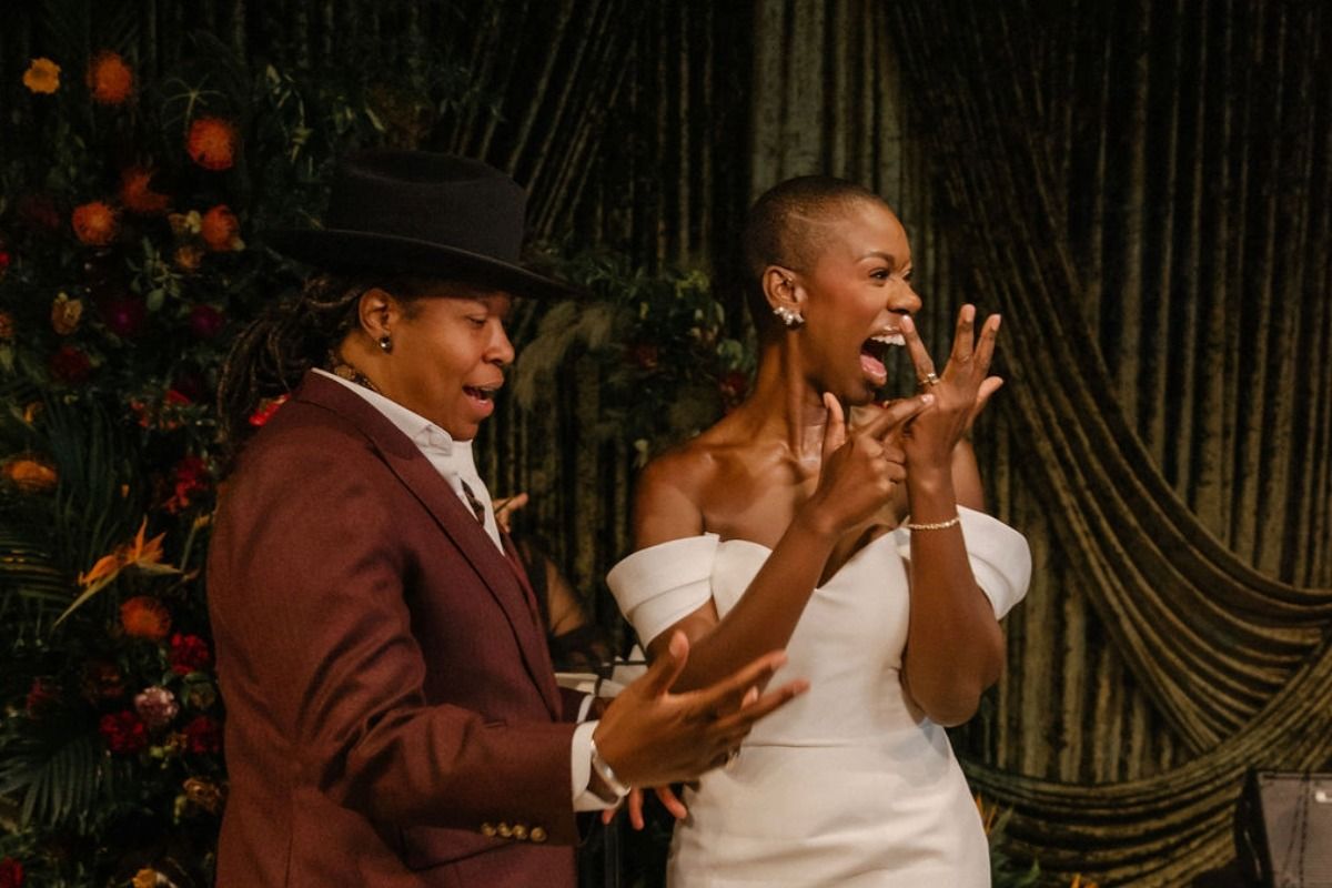Two people celebrate an engagement, one in a dark suit and hat, the other in a white dress, admiring a ring with joy.