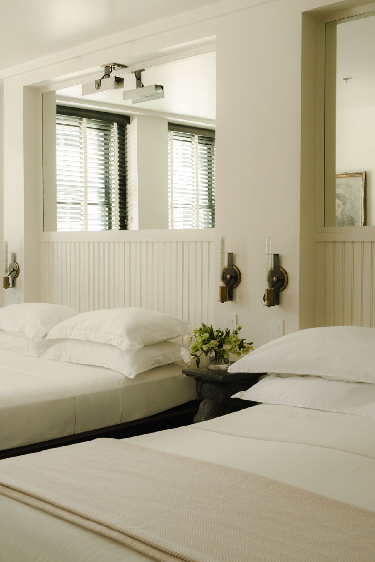 Elegant bedroom with two neatly made beds, white linens, wall sconces, a floral arrangement on the nightstand, and sunlight filtering through blinds.