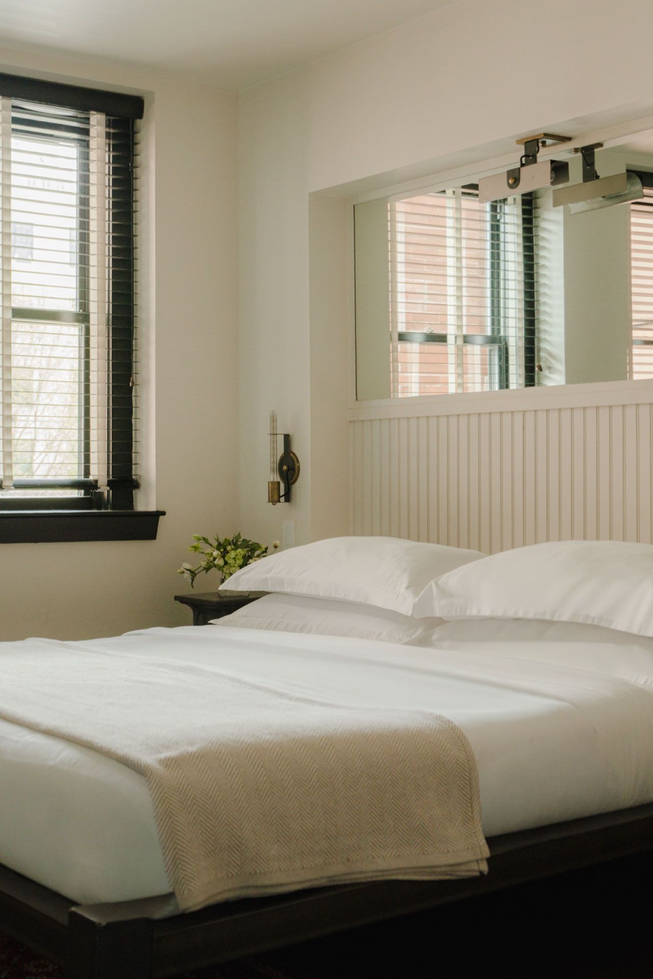 A cozy bedroom with a neatly made bed, white bedding, and a soft blanket, next to a window with blinds and a small plant on a side table.