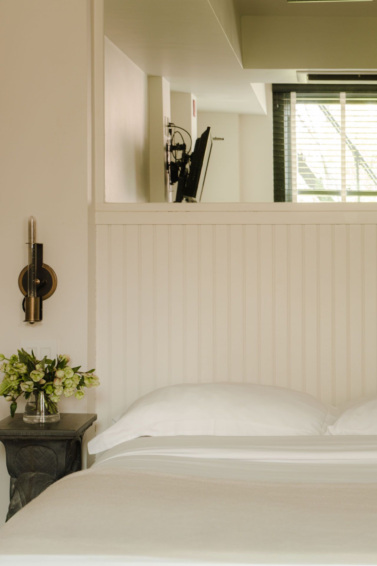 Cozy bedroom with a neatly made bed, white linens, and a bedside table with a vase of green flowers against a light-colored wall.