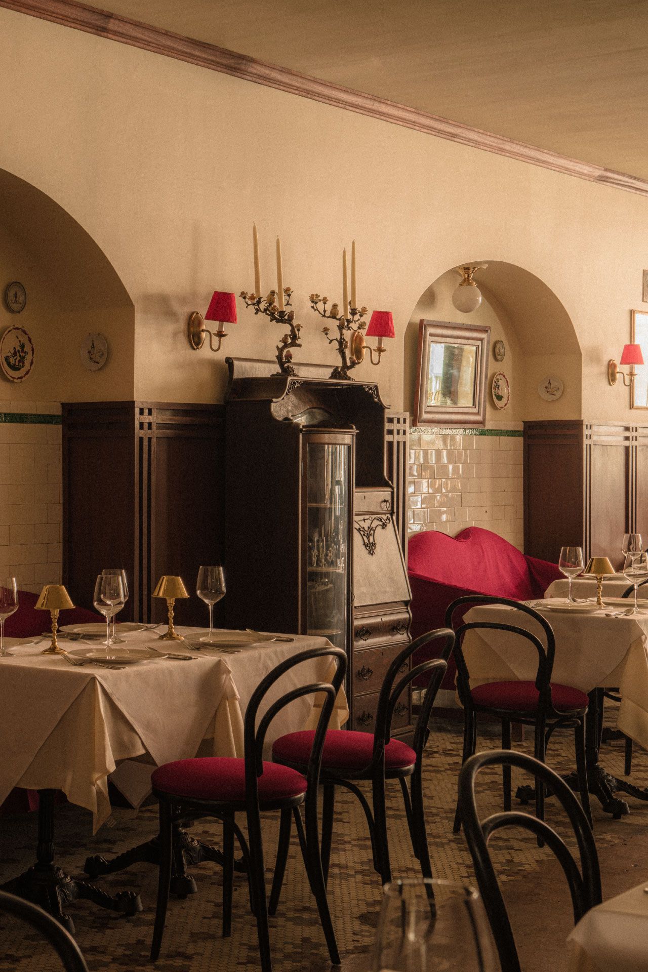Cozy vintage restaurant with arched nooks, white tablecloths, red cushioned chairs, soft lighting, and elegant decor on cream walls.