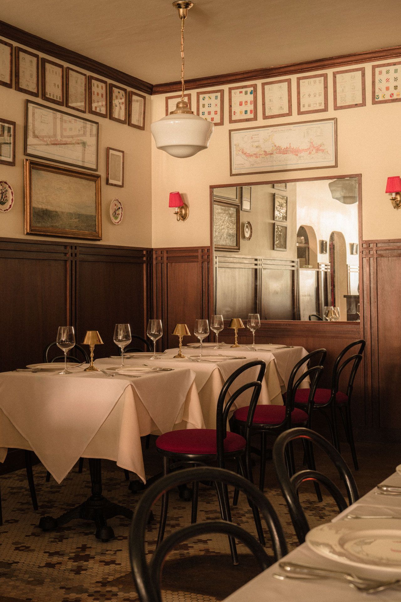 Elegant restaurant interior with tables set with wine glasses, white tablecloths, wooden chairs, framed art on wood-paneled walls, and a large mirror.