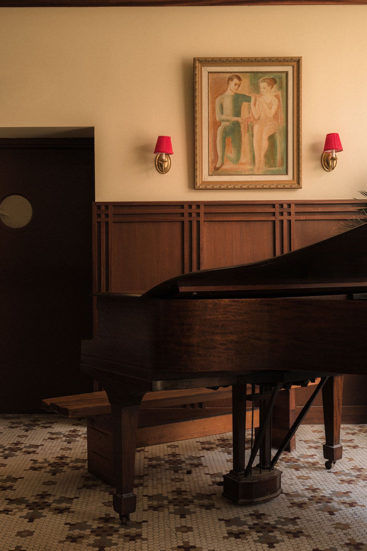 A grand piano in a warmly lit room with patterned tile flooring, wooden paneling, red wall sconces, and a painting of two figures above.