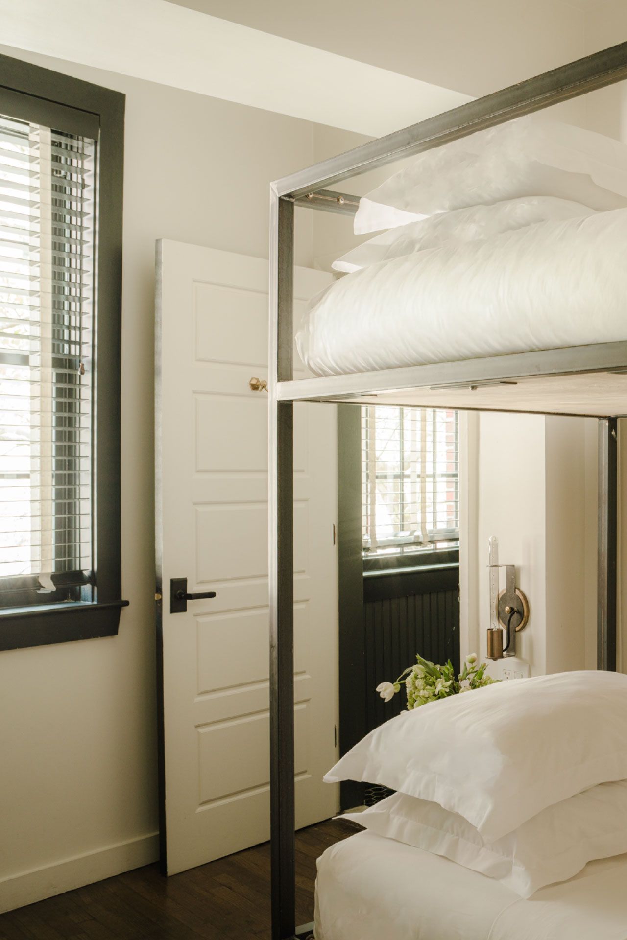 Minimalist bedroom with metal-frame bunk bed, white linens, open door, and window with blinds. Brown wooden floor and small bouquet on the side.