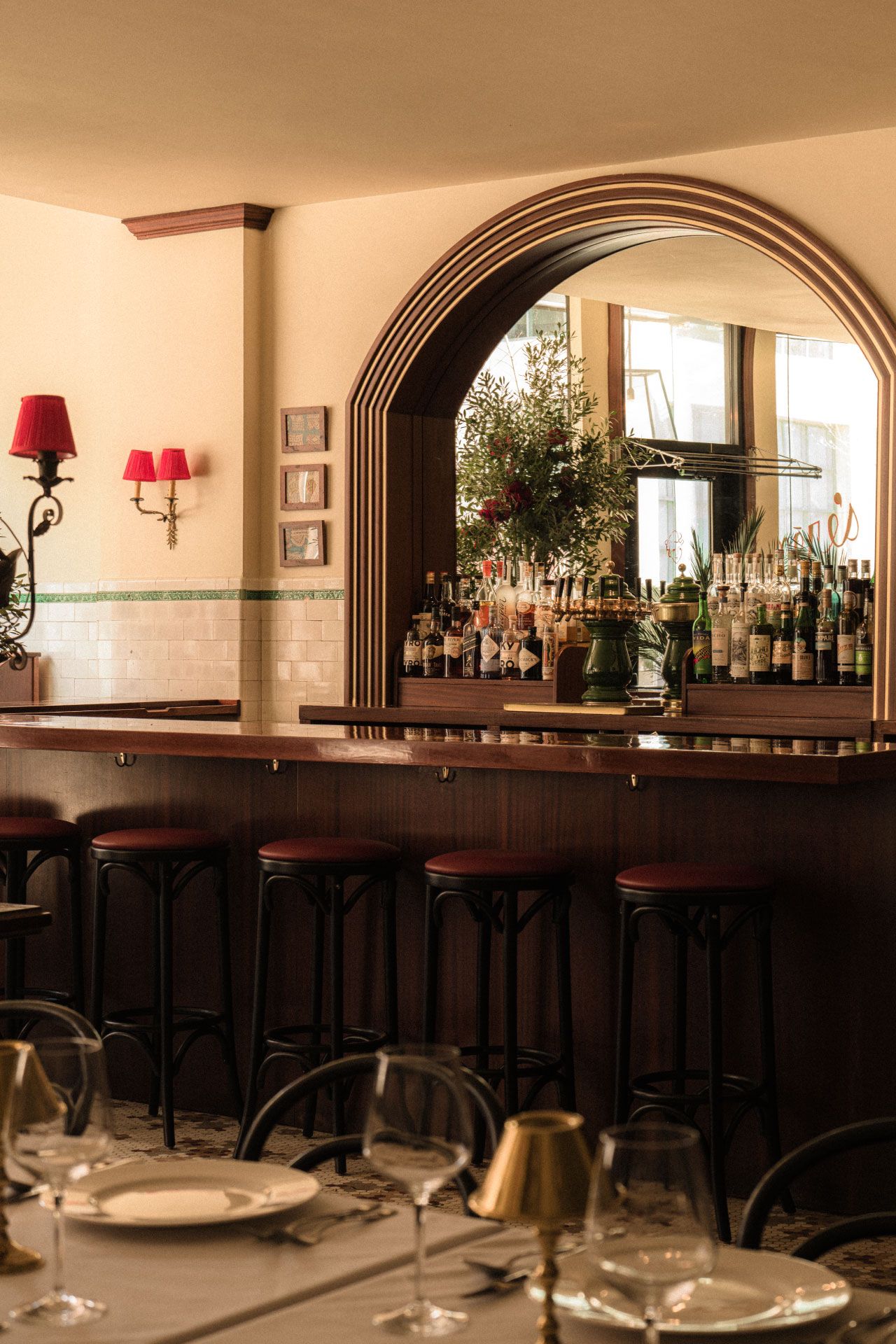 Cozy bar with high stools, a wooden counter, a mirror, and a variety of bottles. Red wall lamps and a set dining table are visible.