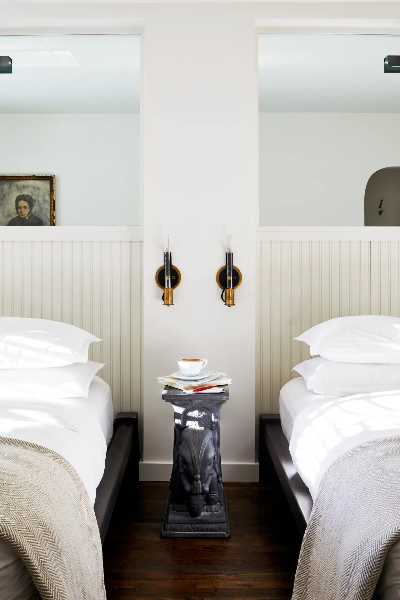 A cozy bedroom with two single beds, a vintage side table holding a teacup, and wall sconces. A portrait hangs on the wall.