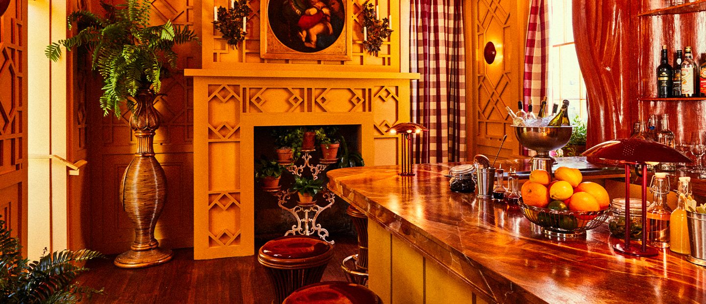 Warm, cozy room with a wooden bar, stools, fruit, plants, and a fireplace. Decor includes a painting above the mantel and plaid curtains.