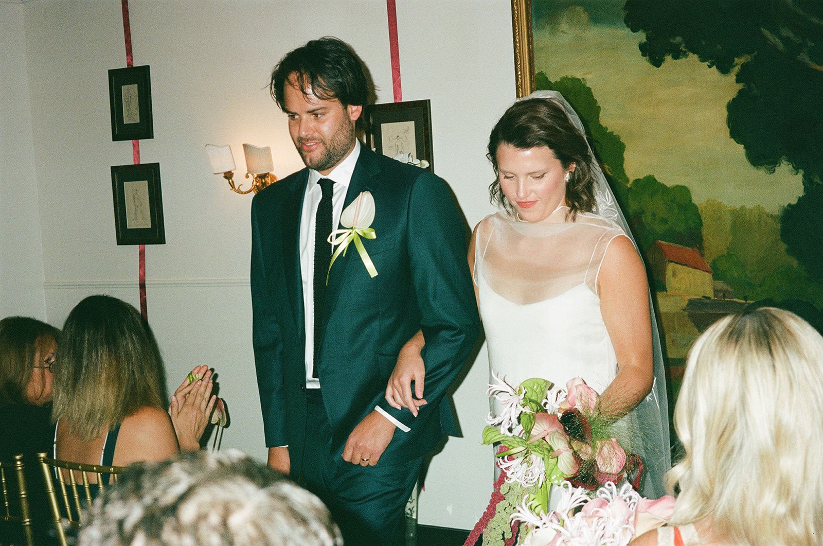 Bride and groom walking arm in arm at a wedding ceremony, with guests seated nearby. Bride holds a bouquet and wears a veil.