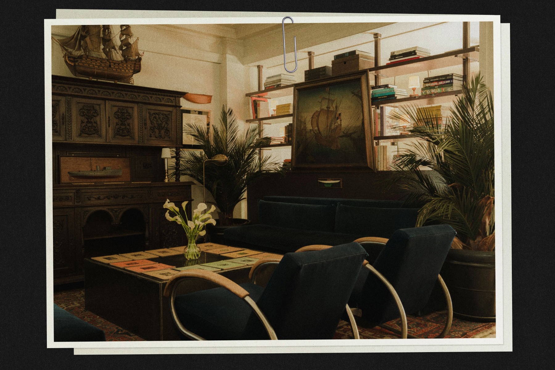 Cozy vintage living room with dark wood furniture, ship model, framed painting, bookshelves, potted plants, and a vase of flowers on a table.