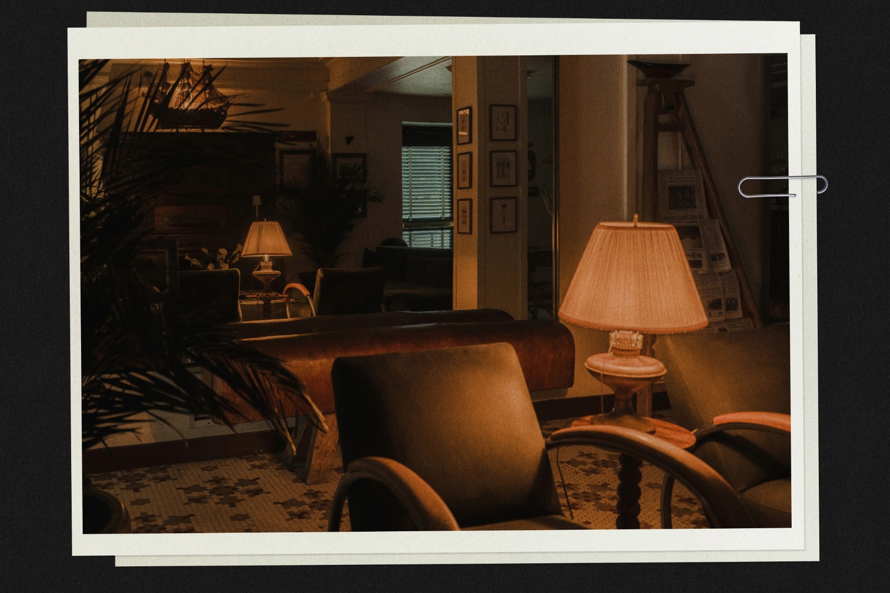 Cozy, dimly lit room with brown leather chairs, a warm table lamp, and framed pictures on the walls. A window with closed blinds in the background.