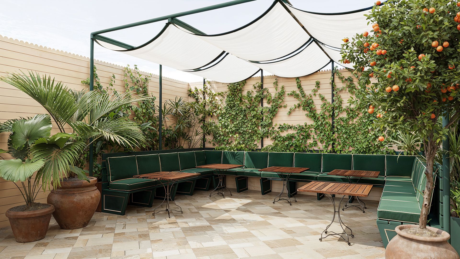 Outdoor patio with green cushioned seating, wood tables, potted plants, ivy-covered walls, and an orange tree under a white canopy.