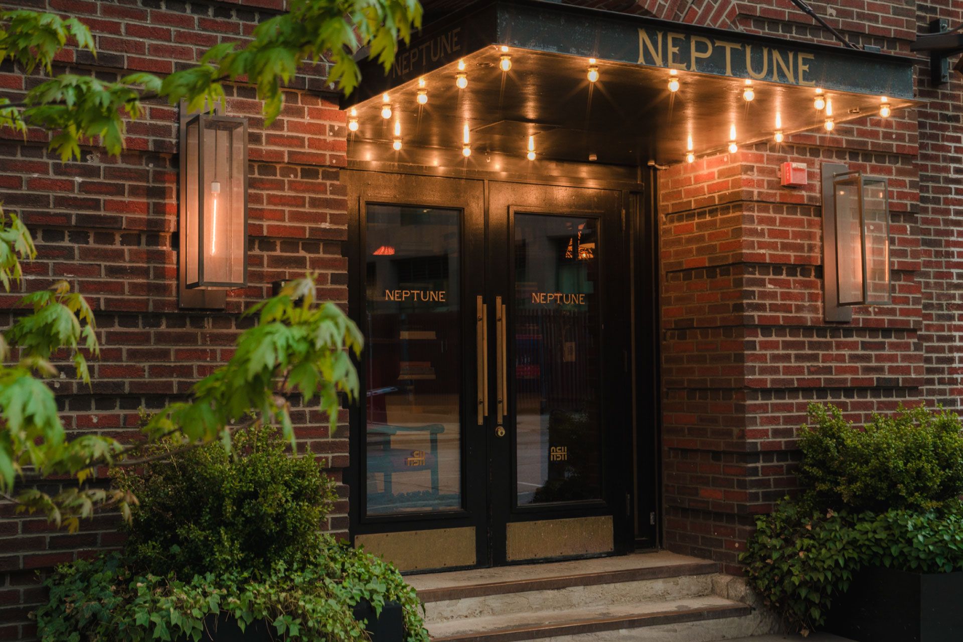 Entrance to "Neptune" with illuminated marquee and brick walls, surrounded by greenery, under warm evening lights.