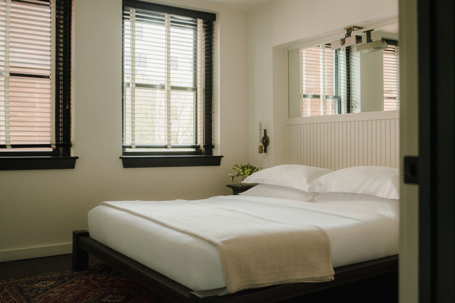 Sunlit bedroom with a neatly made double bed, white bedding, and two windows with blinds. A small side table has a vase with flowers.
