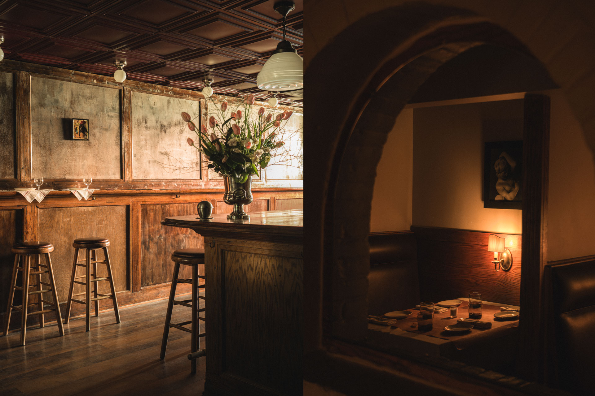 Cozy bar interior with wooden stools, a flower arrangement on the counter, and a dimly lit booth with a table set for dining.