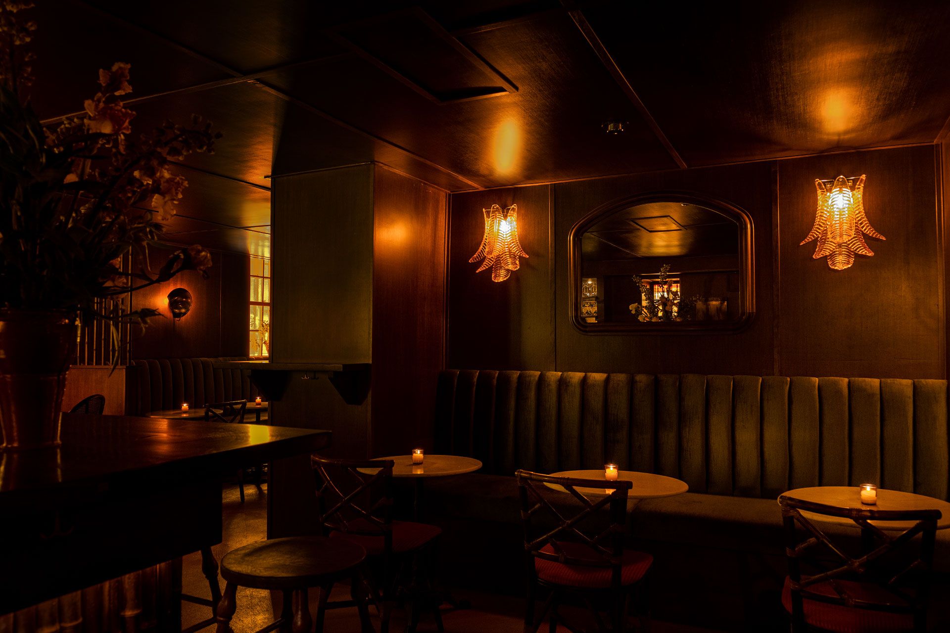 Dimly lit, cozy bar with plush seating, round tables, warm lighting from wall sconces, and a decorative mirror reflecting ambient light.