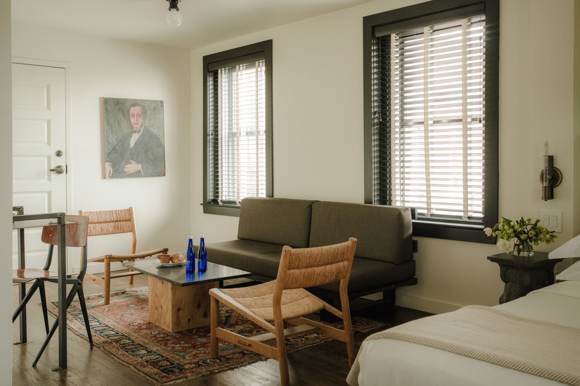 A cozy living room with a grey sofa, wooden chairs, a coffee table with blue bottles, a portrait on the wall, and natural light from the windows.