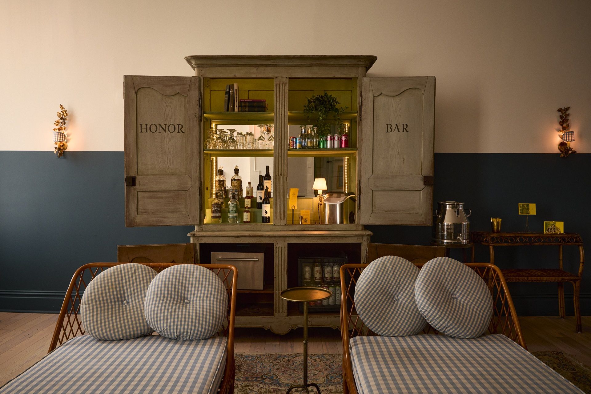 A cozy home bar with open wooden cabinet labeled "Honor Bar," displaying bottles, glasses, and cans. Two wicker chairs with checkered cushions in front.