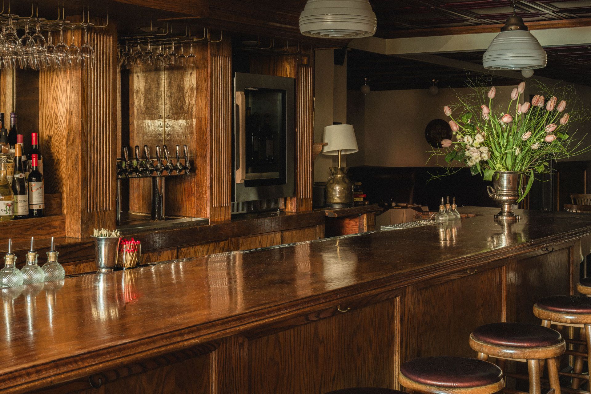 A dimly lit bar with wooden counter, stools, wine bottles, and a vase of pink flowers. Hanging lights create a warm ambiance.