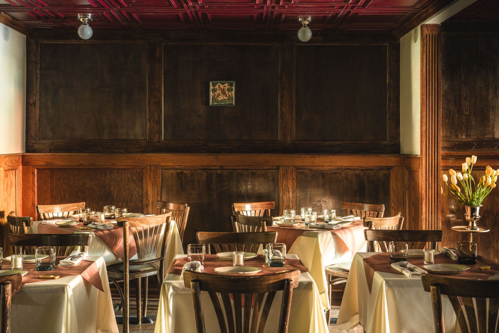 Cozy restaurant with wooden walls, tables and chairs, white tablecloths, set for dining. A vase with tulips decorates the corner.