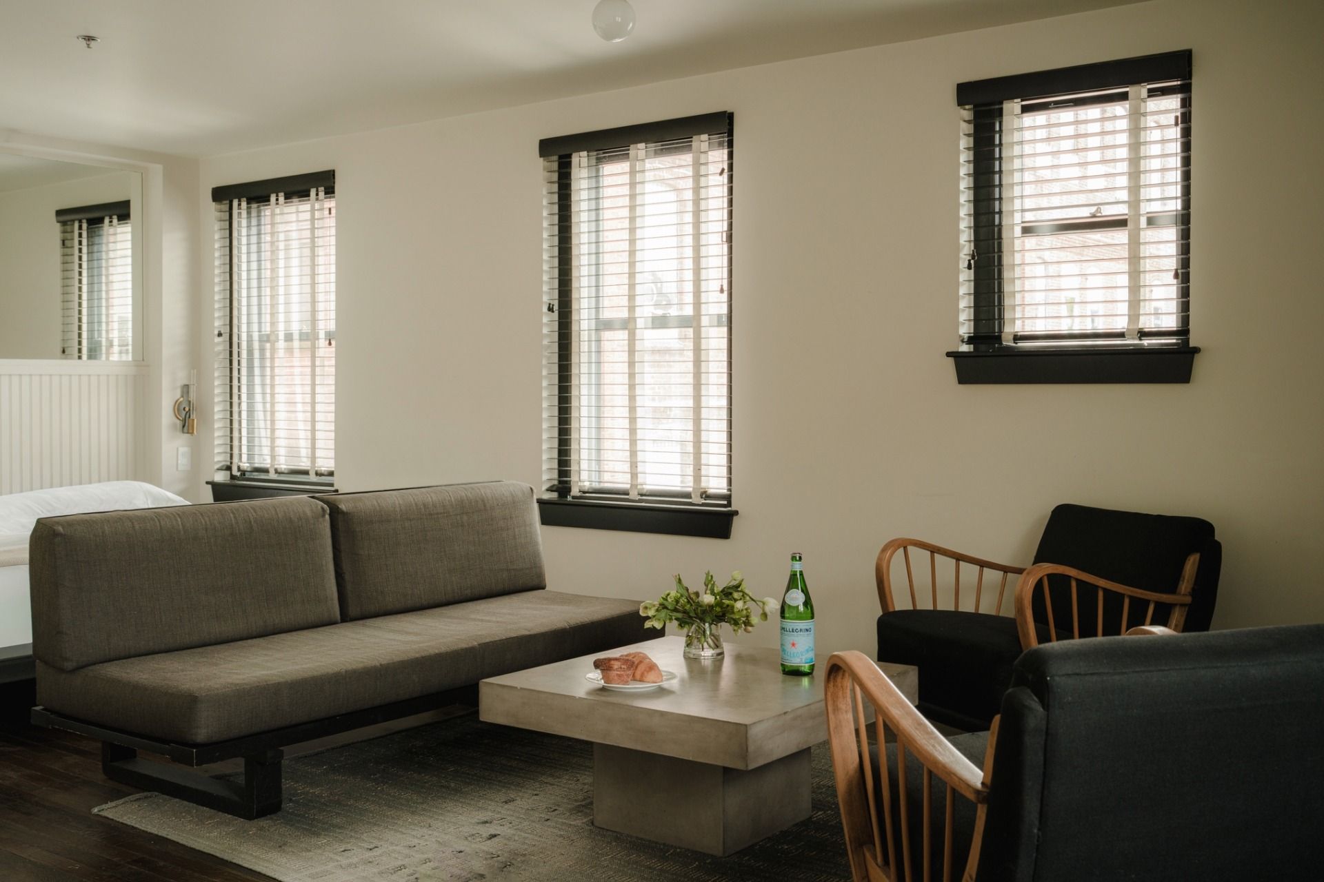 Minimalist living room with a gray sofa, wooden chairs, concrete coffee table, and bright windows. A bottle, bread, and flowers on the table.