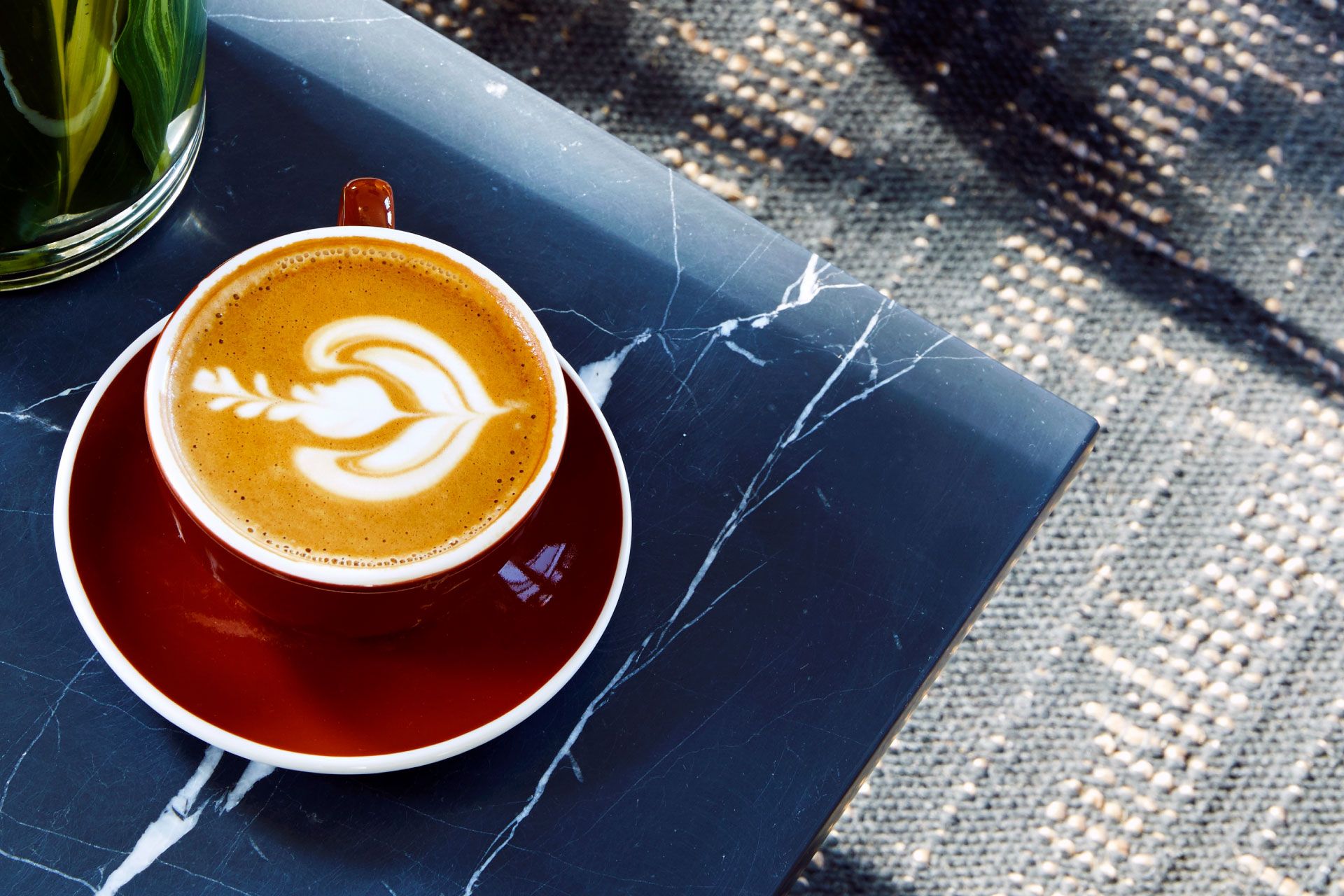 A cappuccino with intricate latte art in a maroon cup and saucer on a dark marble table, with dappled sunlight on a textured floor.