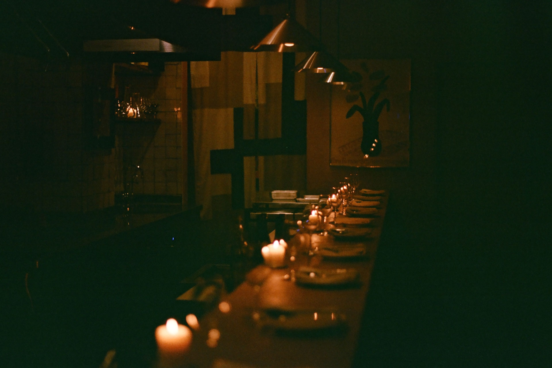 Dimly lit restaurant interior with a long table set with plates, glasses, and candles under hanging lights.