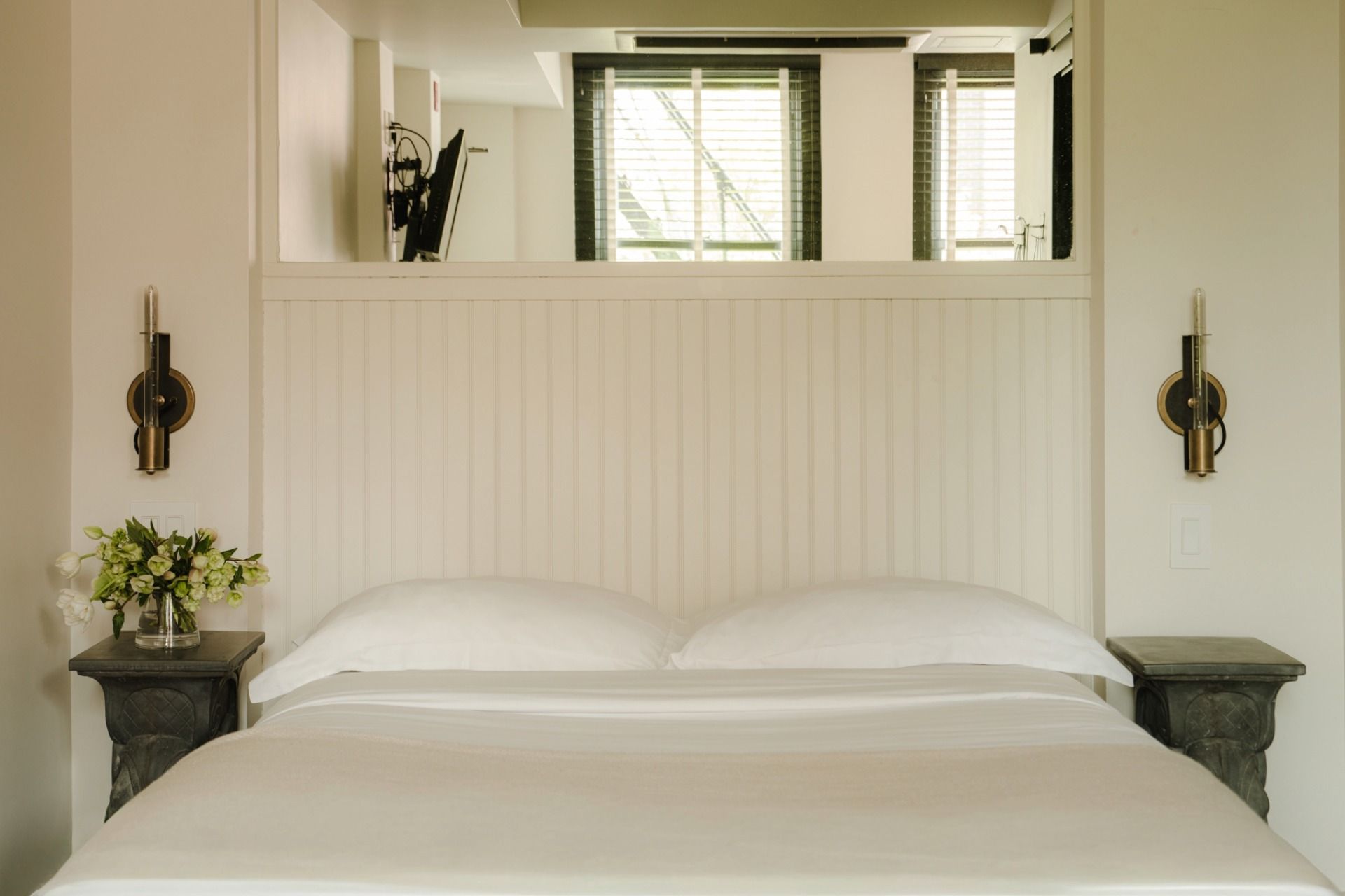 A neatly made bed with white linens, flanked by two sconces. A small table with a vase of flowers is on the left, near a window with blinds.