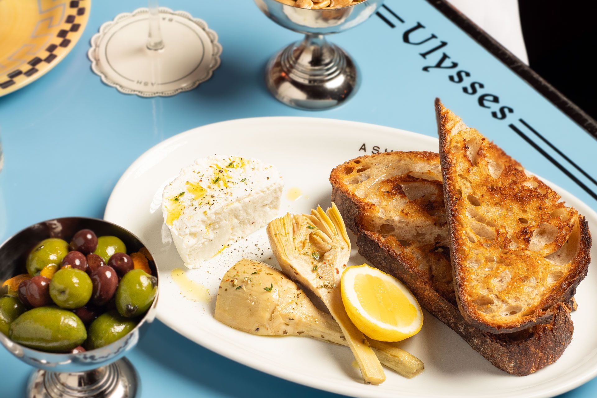 Plate with grilled bread, feta cheese, artichokes, lemon wedge, and herbs. Side of mixed olives on a blue table with "Ulysses" text.