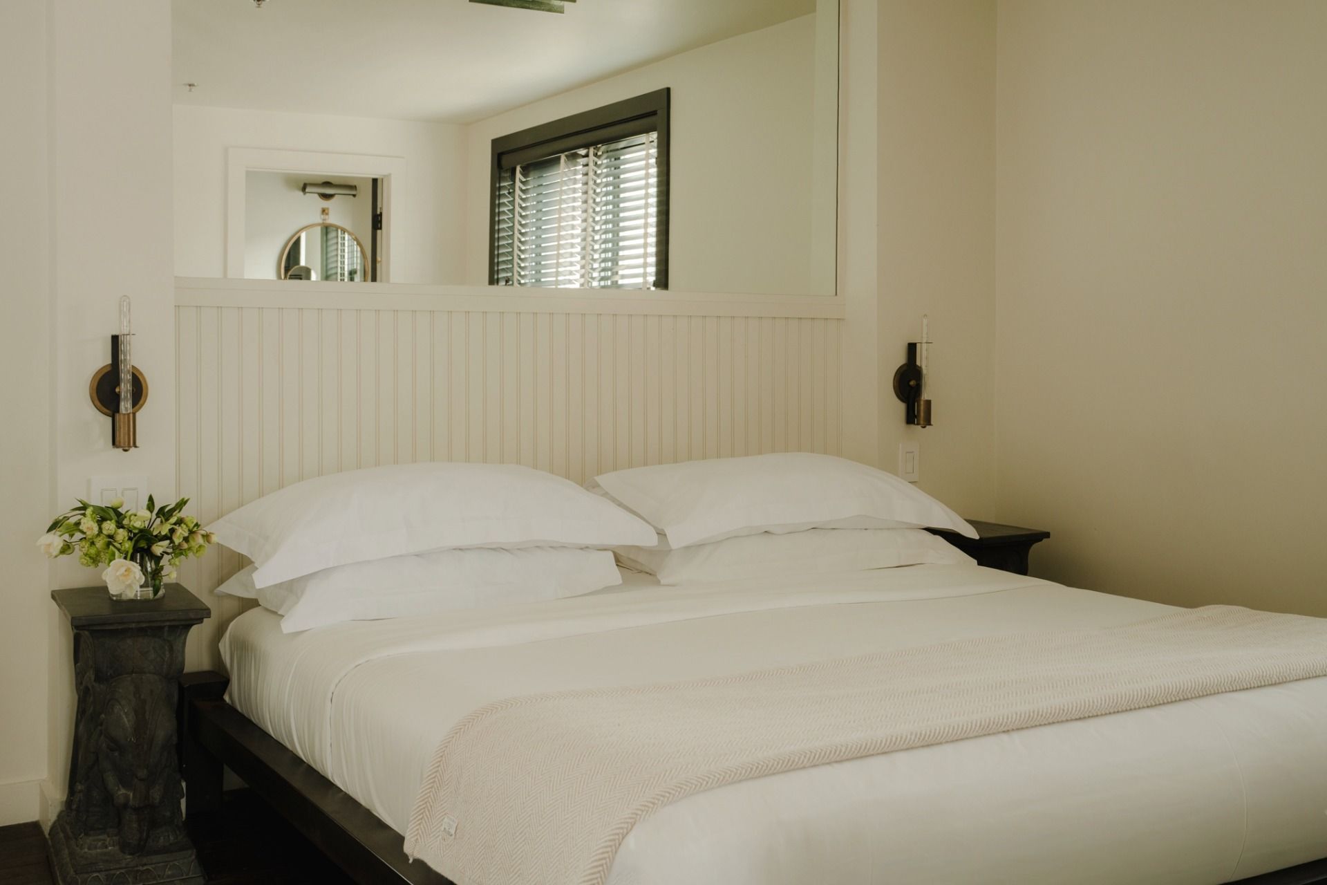 A neatly made bed with white linens and a beige throw, flanked by a decorative table with fresh flowers, in a minimalist room with a window and a mirror.