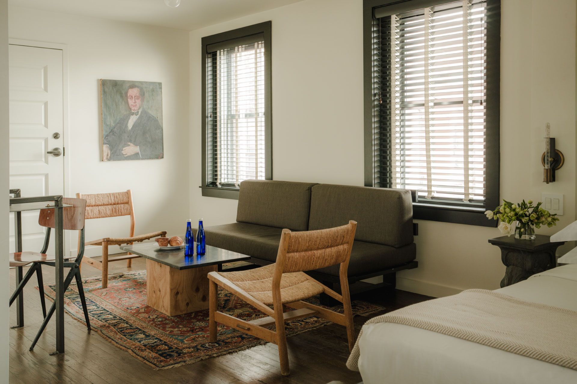 Cozy living room with a gray sofa, wooden chairs, a coffee table, a rug, and a portrait on the wall. Sunlight filters through blinds.