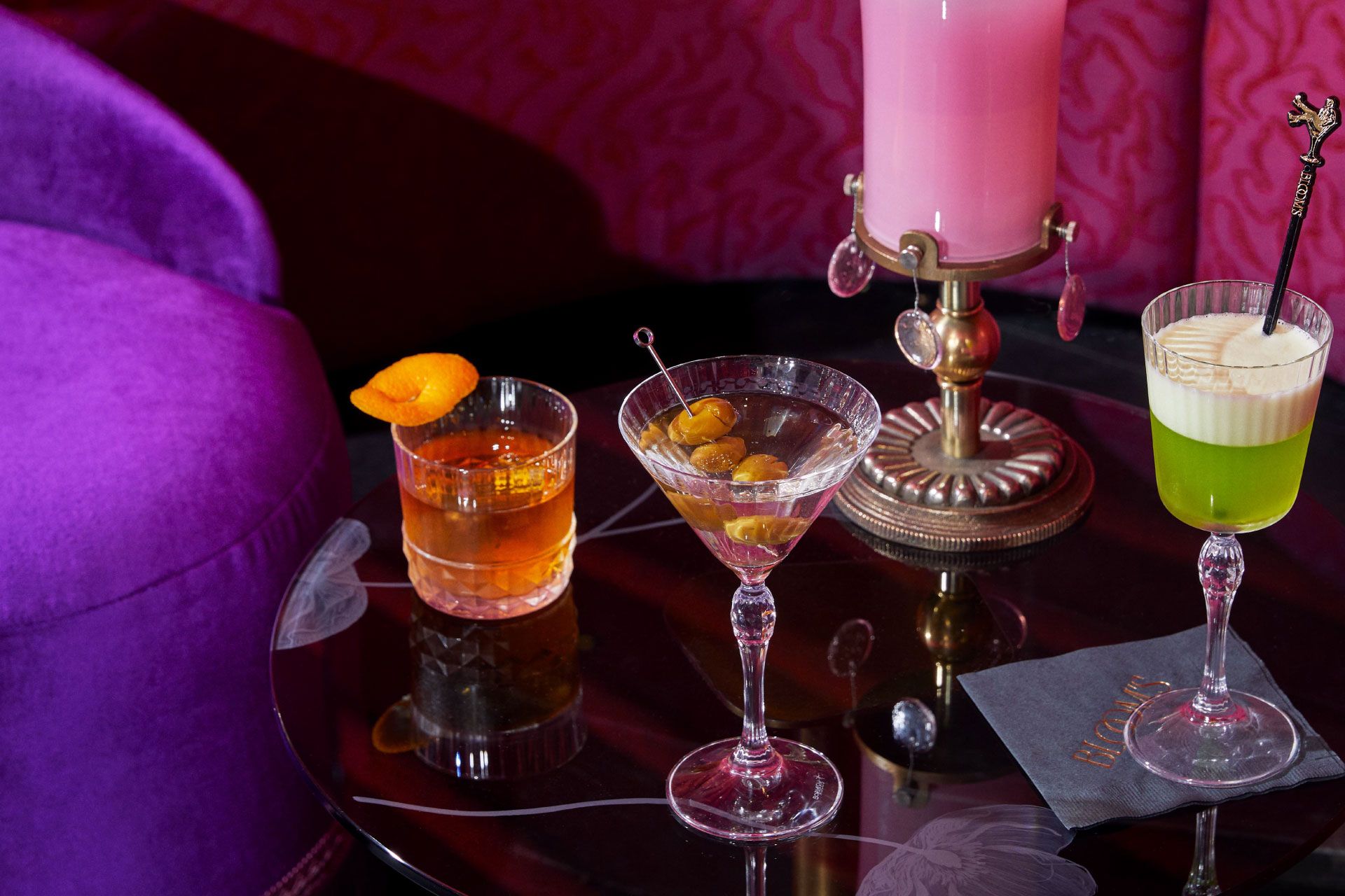 Three cocktails on a glossy table: whiskey with orange garnish, classic martini, and a green drink with foam; plush purple seating in the background.