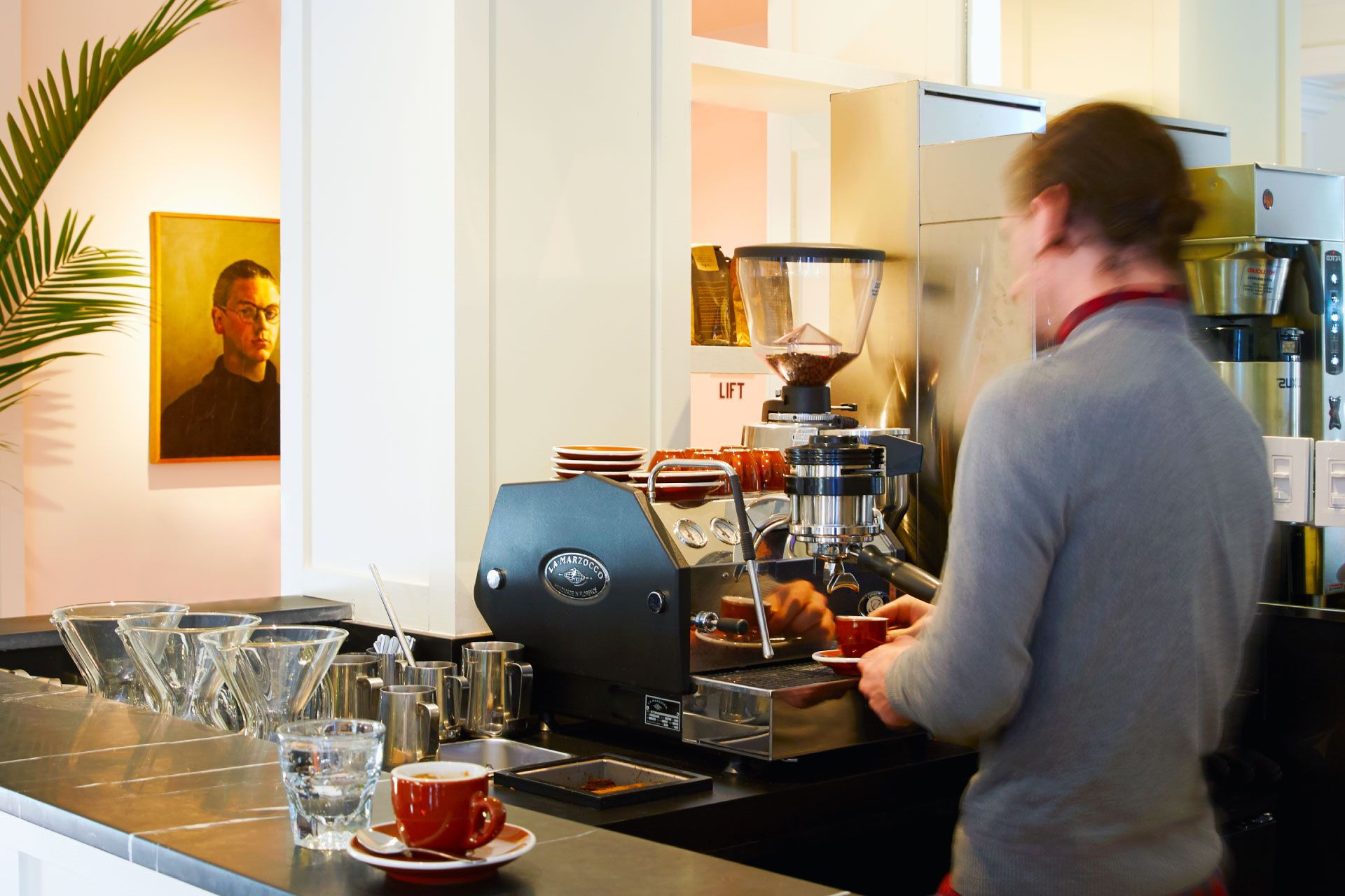 Barista making coffee at an espresso machine, with a small plant and portrait in the background.