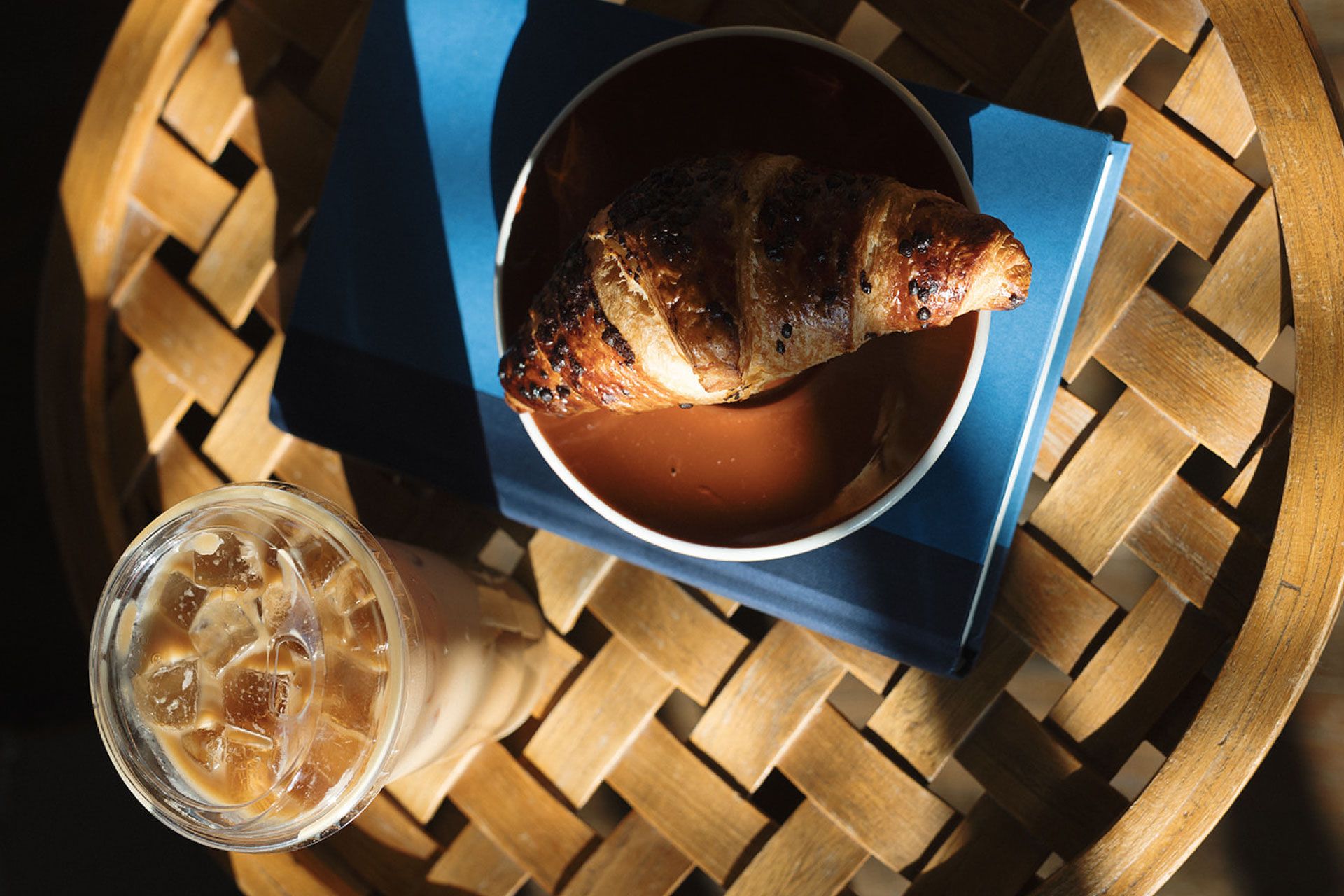 A croissant on a bowl atop a blue book, next to an iced coffee on a woven wooden table with warm sunlight.