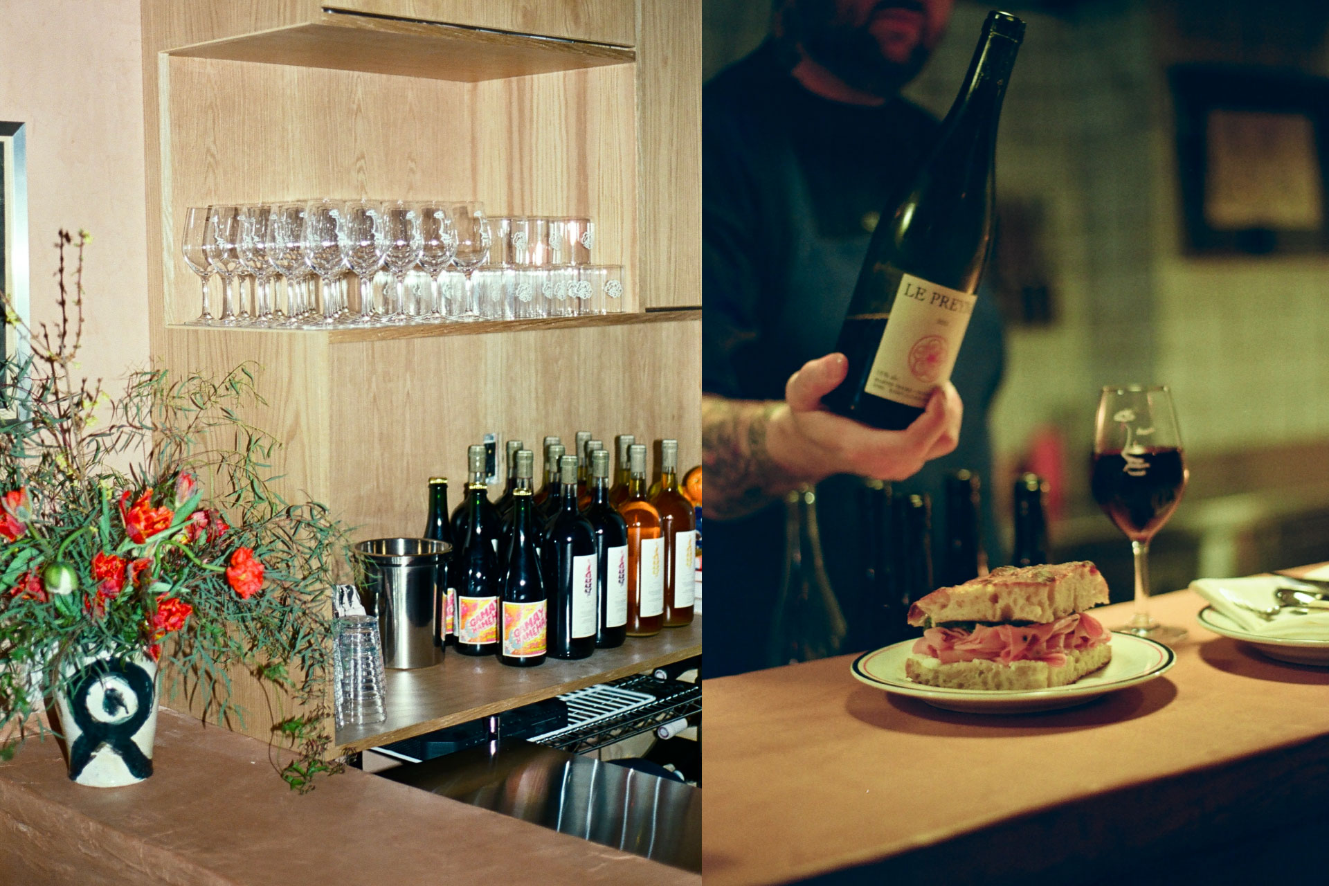 Wooden bar with wine glasses and bottles, a person holding wine, and a plate with a sandwich and glass of red wine on the counter.
