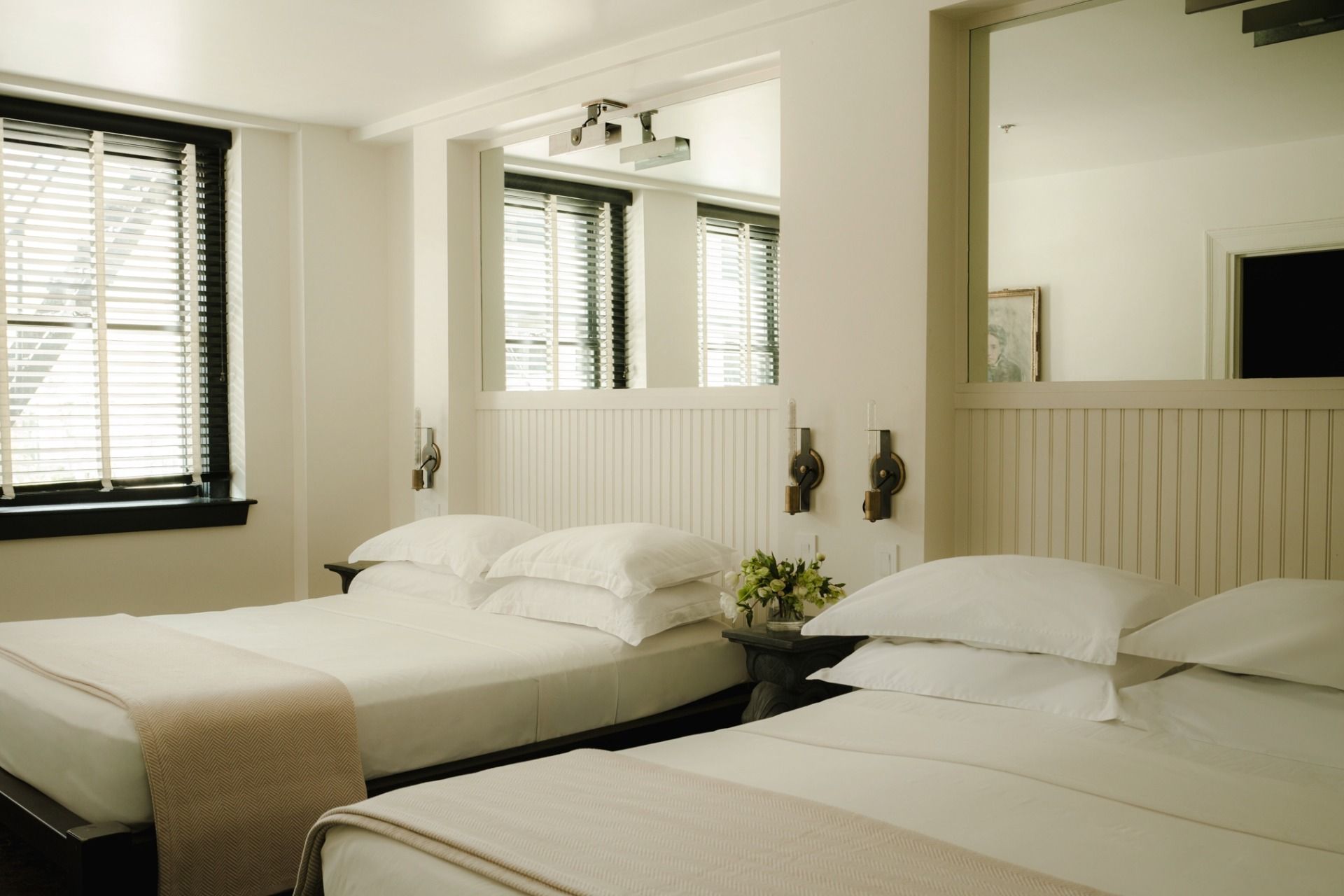 A bright hotel room with two neatly made beds, each with white linens and beige throws, large windows with blinds, and floral arrangements on a side table.
