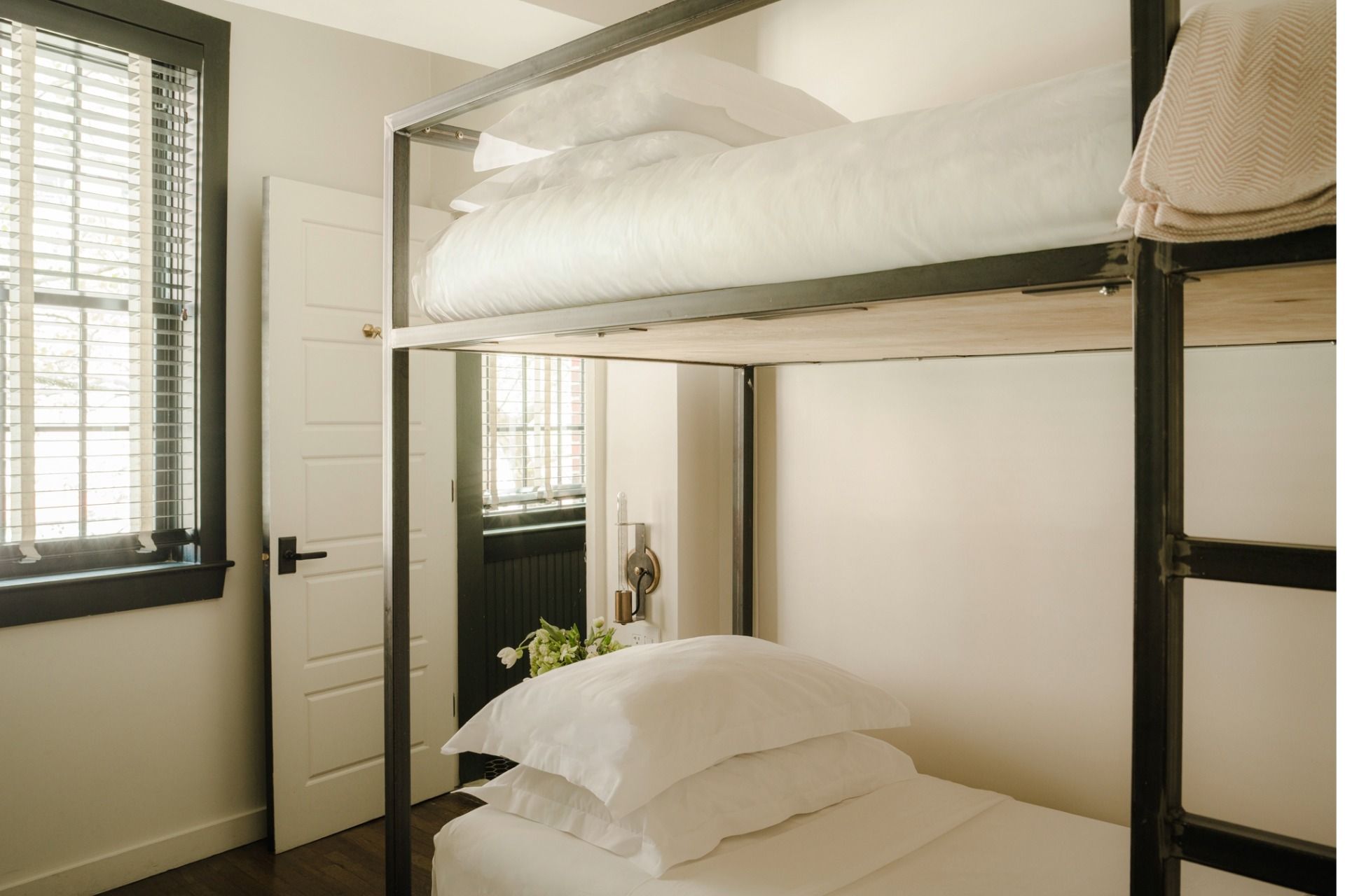 A minimalist bedroom with a metal-framed bunk bed, white linens, a window with blinds, and a partially open door. A stack of towels rests on the top bunk.