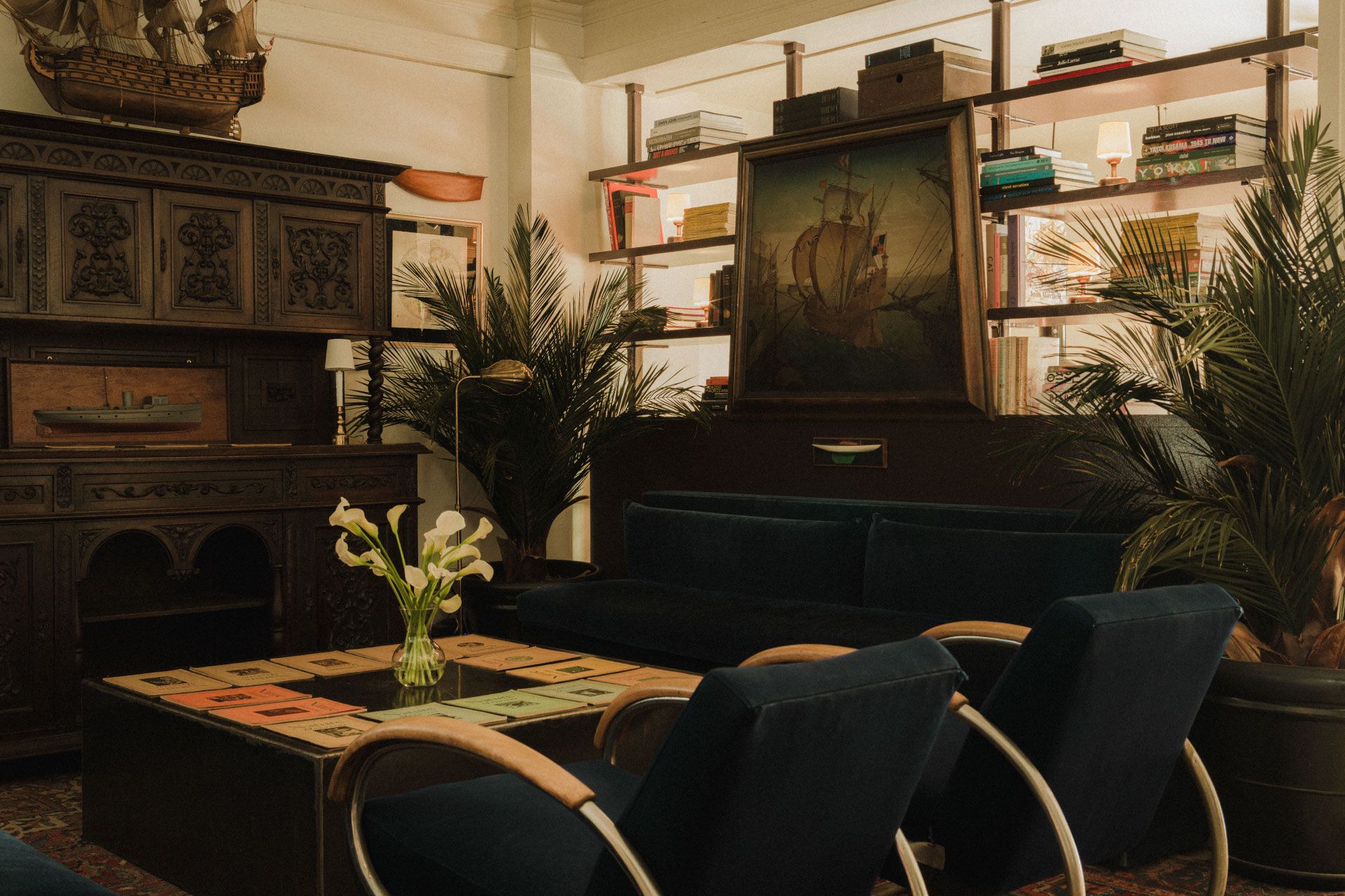 Cozy vintage room with velvet chairs, a wooden cabinet, ship painting, plants, and bookshelves. A vase with white calla lilies on the table.