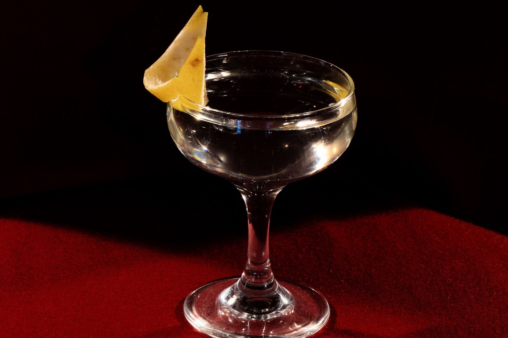 A cocktail glass filled with a clear liquid, garnished with a lemon peel, set on a deep red surface against a dark background.