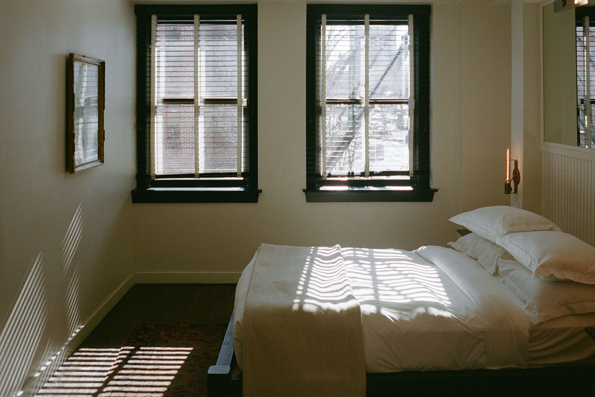 A sunlit bedroom with two windows, a neatly made bed with white linens, and light streaming through blinds, casting shadows on the hardwood floor.