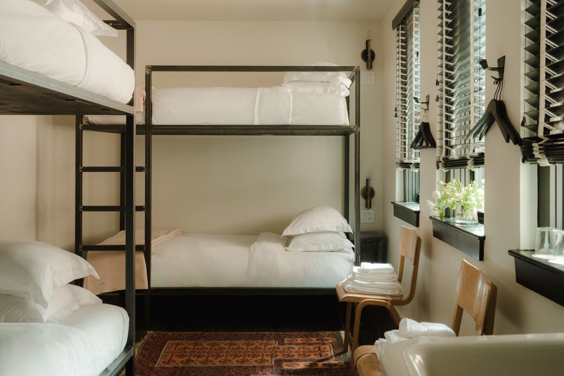 Room with two bunk beds, neat white bedding, a rug, wooden chairs with folded towels, and windows with blinds and hangers.