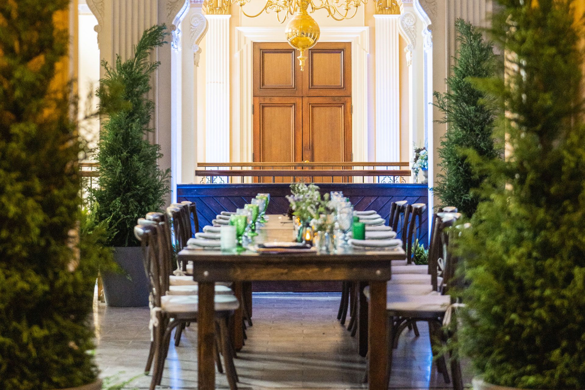 Elegant dining table set with white plates and green glasses, surrounded by greenery, in a grand hall with ornate columns and chandelier.