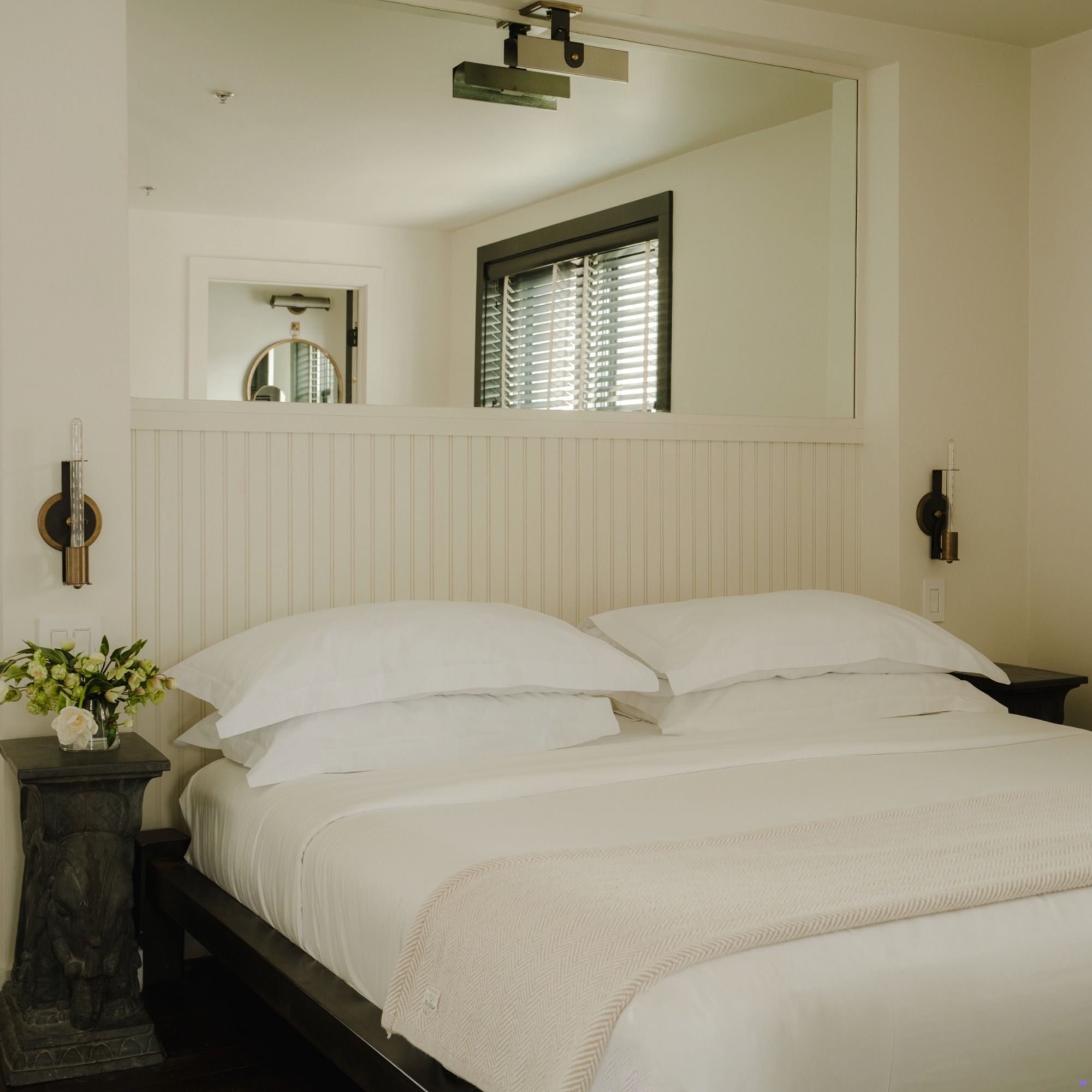 A neatly made bed with white linens, flanked by two bedside tables, a wall mirror above, and a window with blinds in a minimalist bedroom.